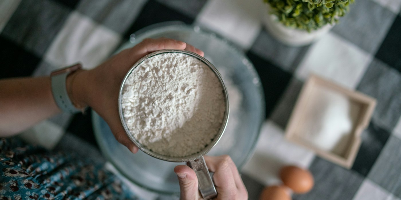 Hands holding a metal measuring cup full of flour over a mixing bowl on a checked tablecloth