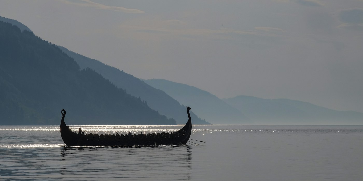 A replica Viking longship with a crew rowing across a still fjord, mountains fading into haze behind it